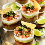 Five glass cups showcase a vibrant 5 Layer Dip, topped with diced tomatoes, olives, and green onions, each garnished with crunchy tortilla chips. Lime wedges and fresh cilantro beautifully adorn the rustic wooden surface.