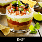 A glass dish presents a vibrant 5 Layer Dip, crowned with olives, diced tomatoes, green onions, and a tortilla chip. Lime wedges and fresh cilantro rest invitingly nearby.