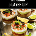 Glass cups brimming with a zesty 5-layer dip, crowned with diced tomatoes, olives, and green onions, are surrounded by crunchy tortilla chips and lime wedges on a wooden surface.