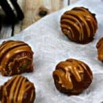 Five chocolate truffles, reminiscent of Peanut Butter Bon Bons, with a drizzle of caramel on top are placed on a crumpled piece of parchment paper, with an Oreo cookie partially visible in the background.