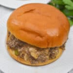 A Philly Cheese Steak Sloppy Joe rests on a white plate, crowned with a glossy bun and packed with seasoned ground beef and onions. In the background, fresh parsley adds a pop of color.