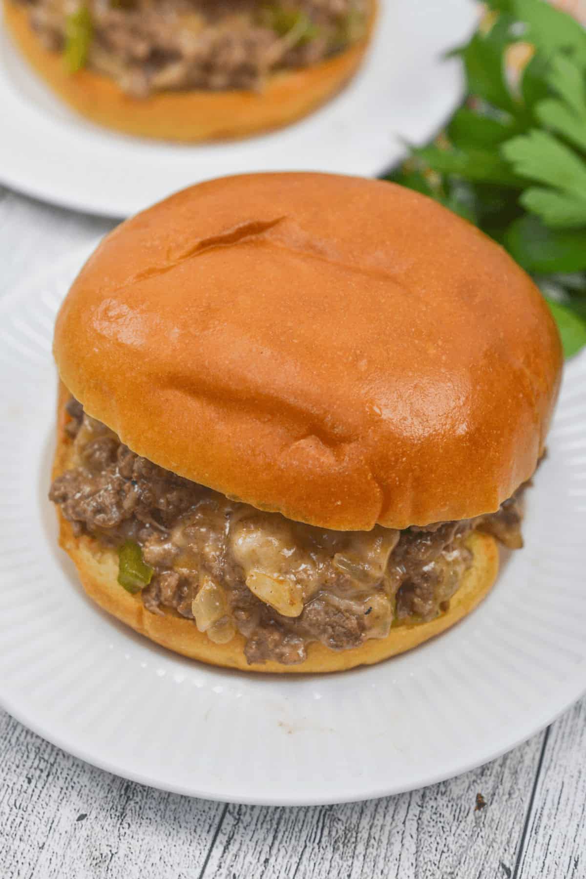 A sandwich rests on a white plate, crowned with a glossy bun and packed with seasoned ground beef and onions. In the background, fresh parsley adds a pop of color.