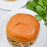 A Philly Cheese Steak Sloppy Joe sandwich rests on a white plate, accompanied by a side of fresh parsley, all set atop a rustic wooden table.