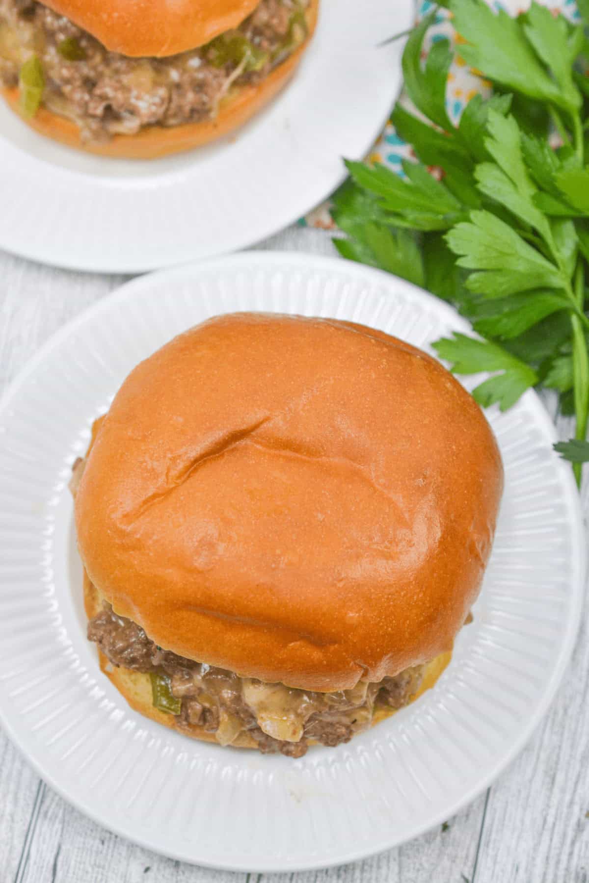 A Philly Cheese Steak Sloppy Joe sandwich rests on a white plate, accompanied by a side of fresh parsley, all set atop a rustic wooden table.