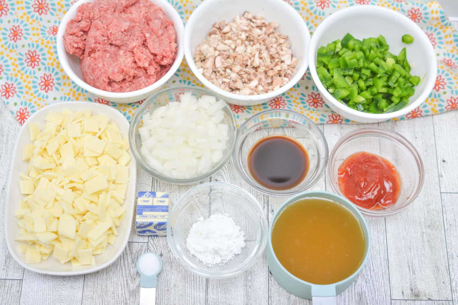 Ingredients for a delightful twist on are displayed on a table: ground meat, diced onions, mushrooms, green bell peppers, sliced cheese, broth, ketchup, Worcestershire sauce, cornstarch, garlic powder, and butter.