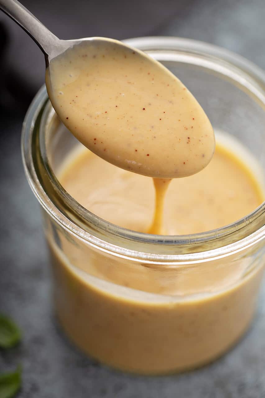 A spoonful of creamy yellow sauce with specks is lifted from a glass jar half-filled with spicy honey mustard goodness.