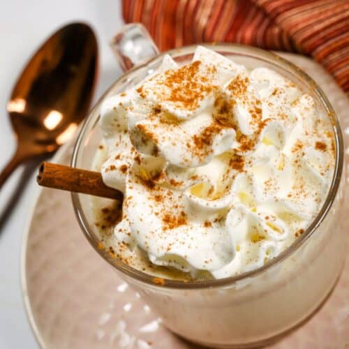 A glass mug filled with White Chocolate Hot Chocolate, topped with whipped cream, ground cinnamon, and a cinnamon stick, sits on a saucer beside a spoon and a striped cloth.