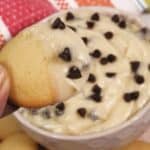 A hand holds a vanilla wafer dipped in rich Chocolate Chip Dip, topped with chocolate chips, with more wafers and a bowl of the delicious dip in the background.