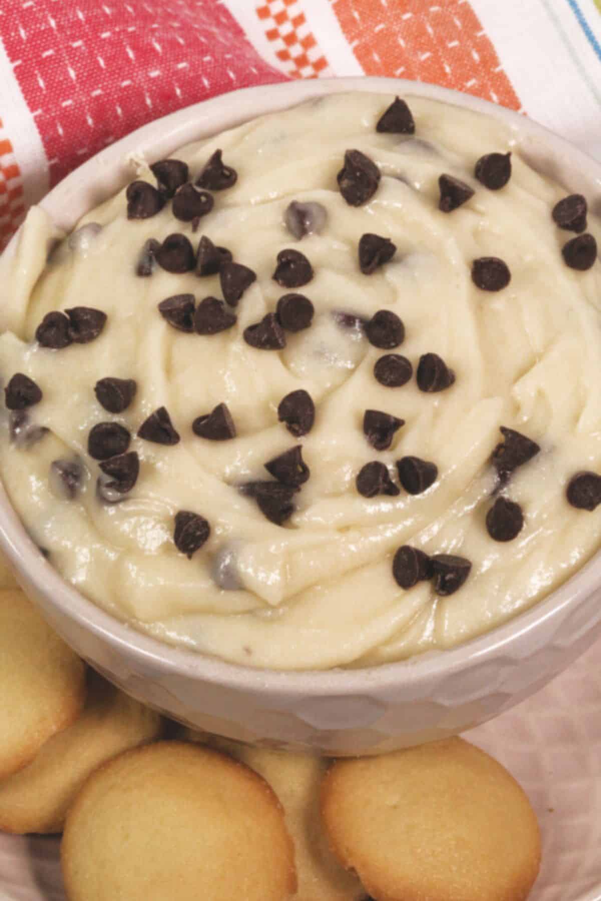 A bowl of creamy treat topped with chocolate chips, surrounded by round vanilla cookies, rests on a patterned cloth.