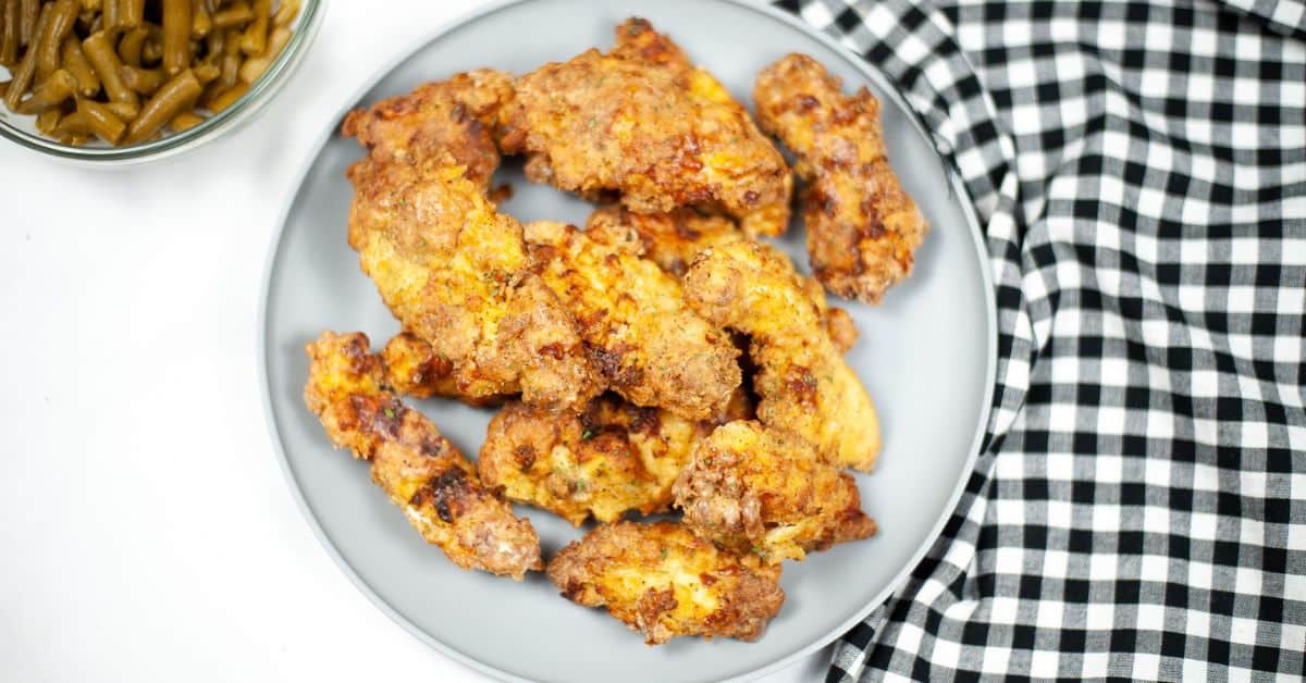 A plate of crispy Air Fryer Chicken Strips is placed next to a black and white checkered cloth, with a bowl of green beans partly visible in the corner.