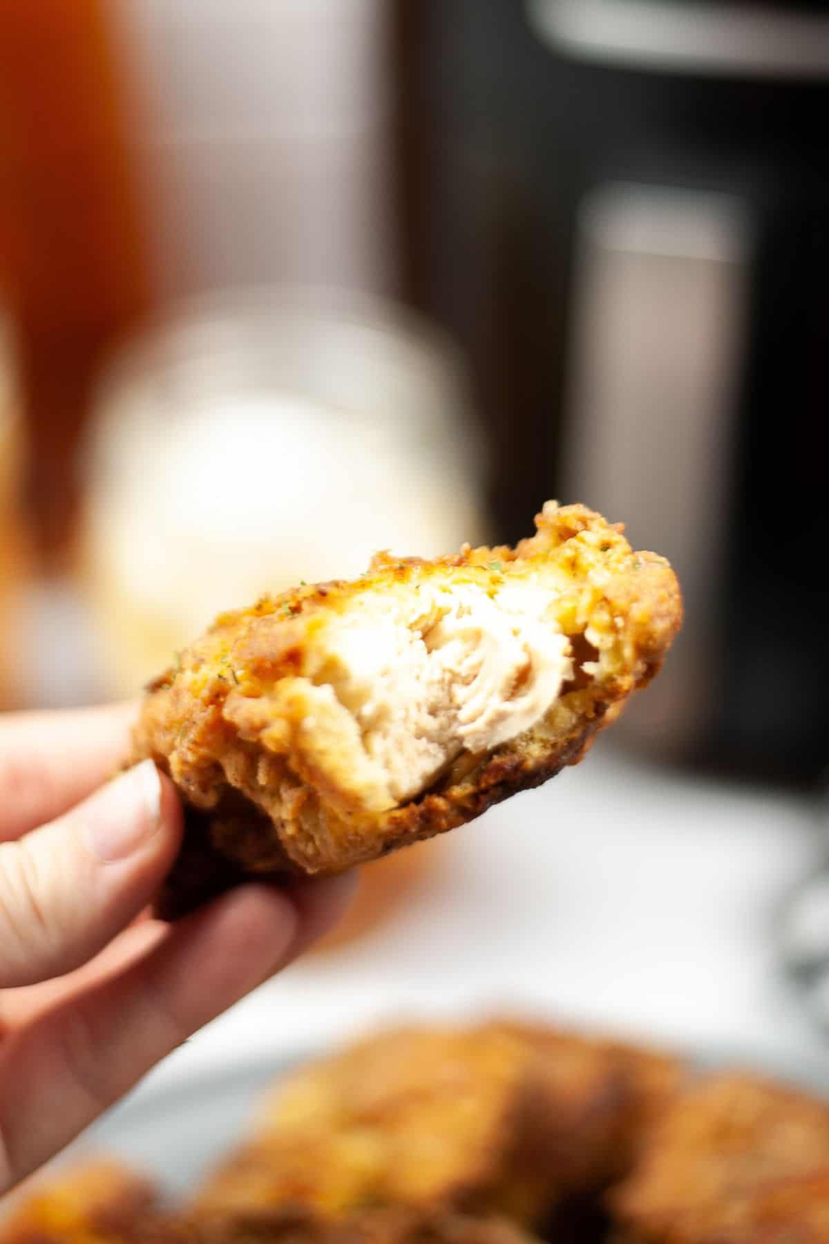 A hand holds a piece of Air Fryer Chicken Strip with a bite taken out, revealing the tender white meat inside.