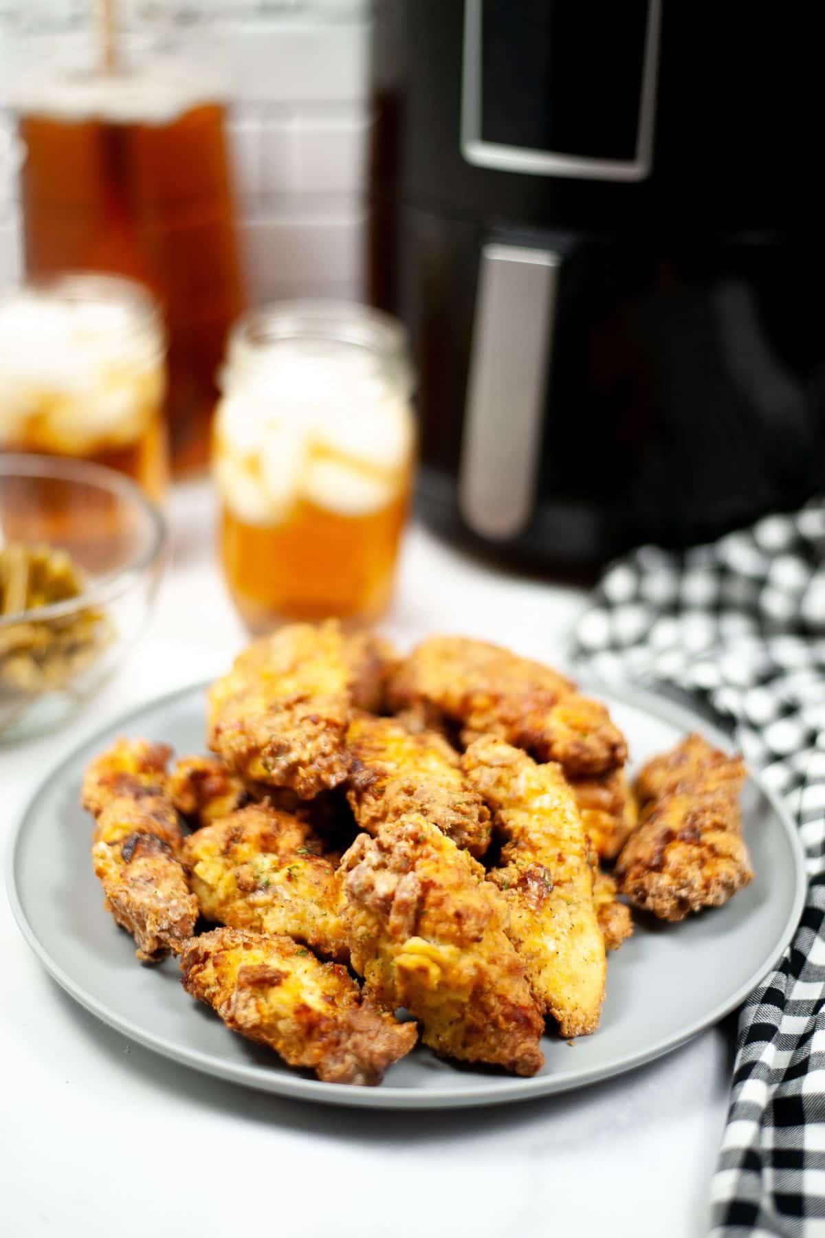 A plate of crispy Air Fryer Chicken Strips sits on a table with iced tea in mason jars and pickles in the background, next to a sleek black air fryer and a checkered cloth.