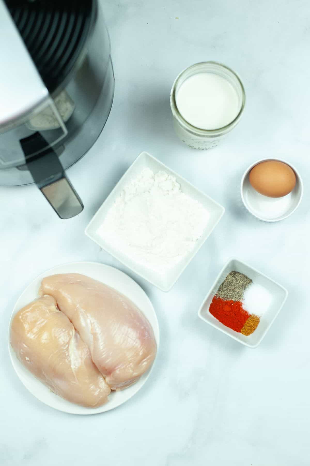 Two raw chicken breasts, flour, spices, an egg, a jar of milk, and an air fryer are arranged on a white surface—everything you need to make delicious Air Fryer Chicken Strips right at home.