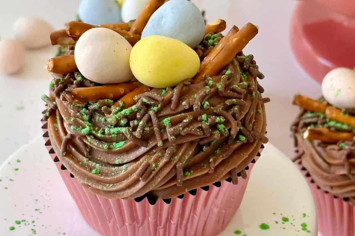 A delightful Easter treat for kids: a cupcake with chocolate frosting, topped with green sprinkles, pretzel sticks, and pastel-colored candy eggs, all nestled in a pink wrapper.
