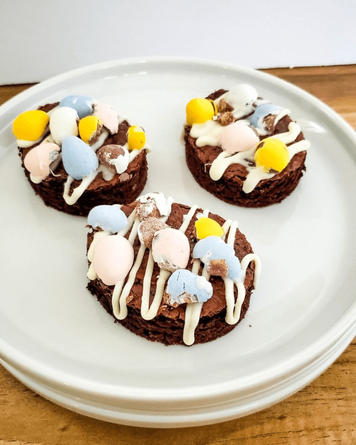 Three chocolate easter brownies topped with pastel Easter candies and white drizzle on a plate.
