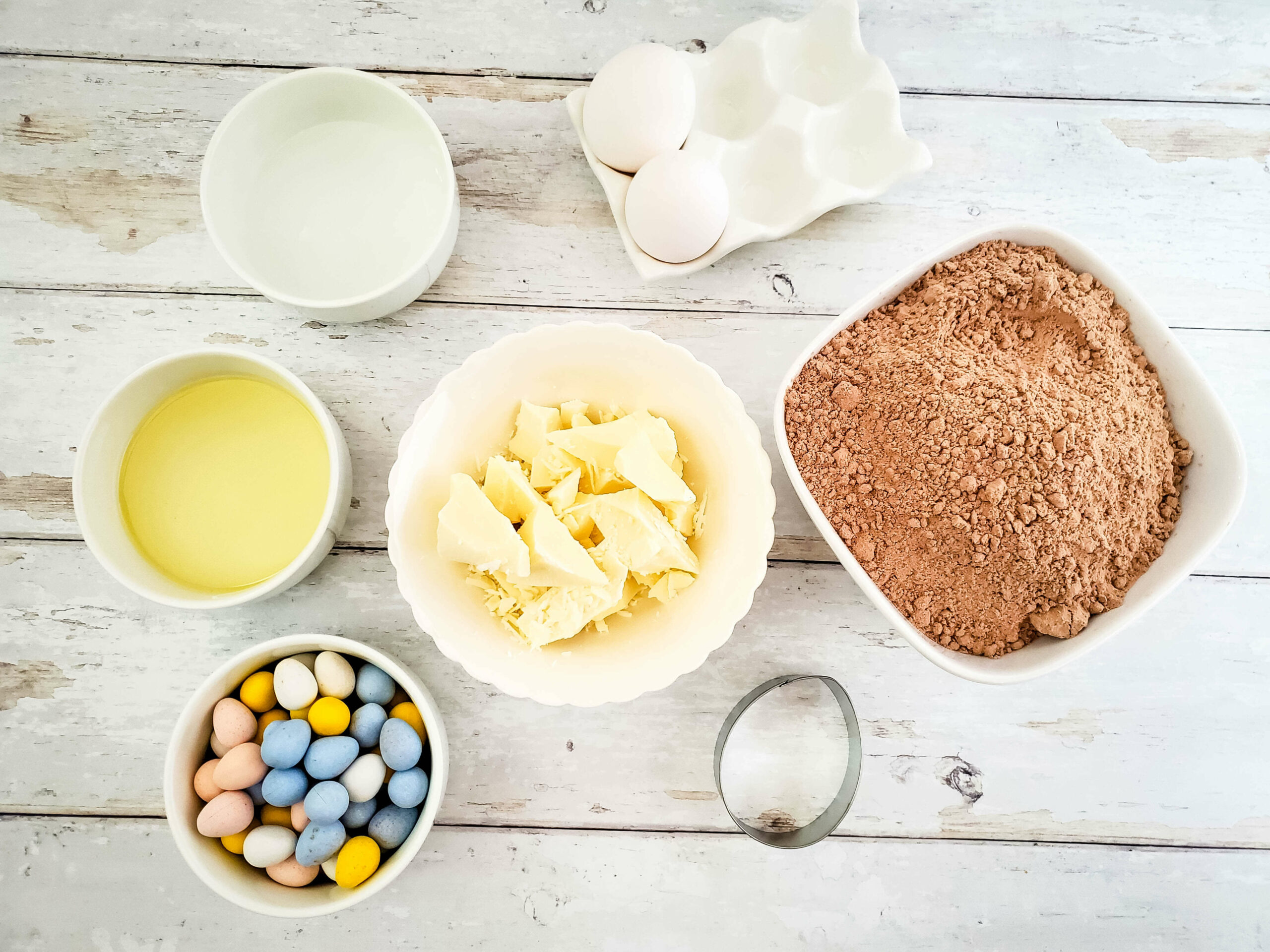 Ingredients for baking Easter brownies arranged on a wooden surface, including eggs, flour, butter, sugar, milk, and candy eggs.