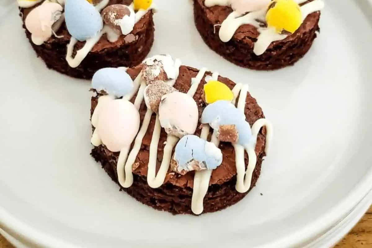 Three round chocolate brownies, topped with pastel-colored candy eggs and a white icing drizzle, make delightful Easter treats for kids. Served on a white plate, these goodies are sure to brighten any child's holiday celebration.