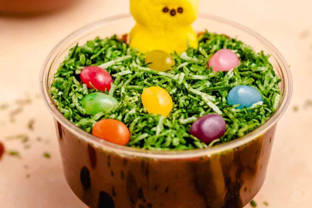 A clear cup filled with crushed chocolate cookies, topped with green coconut flakes, colorful candy eggs, and a yellow marshmallow chick makes for one of the delightful Easter treats for kids.