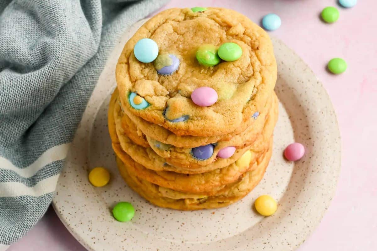 A stack of cookies with colorful candy pieces on a speckled plate, perfect as Easter treats for kids, sits next to a folded cloth.