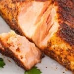 A close-up of a seasoned, cooked jerk salmon fillet with a piece cut off, served on a white plate with parsley garnish.
