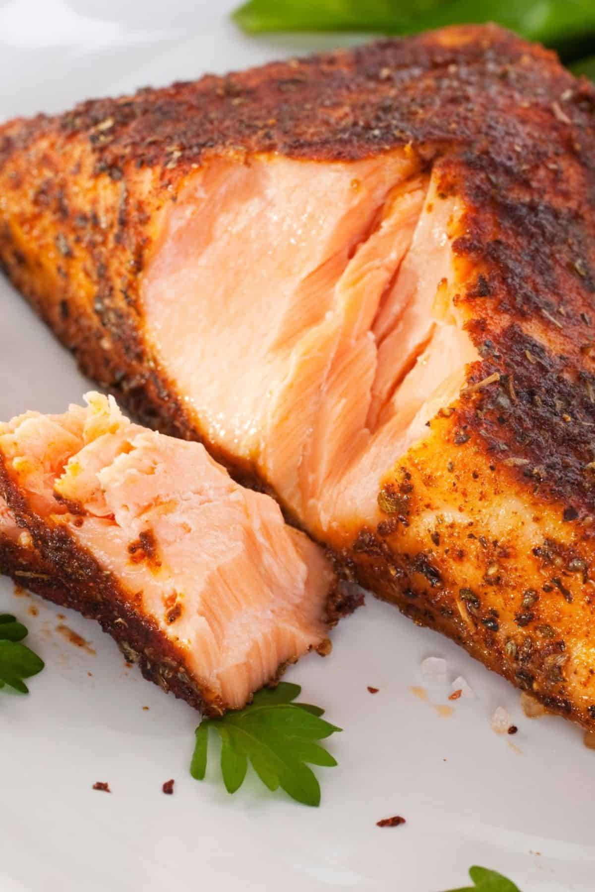 A close-up of a seasoned, cooked jerk salmon fillet with a piece cut off, served on a white plate with parsley garnish.