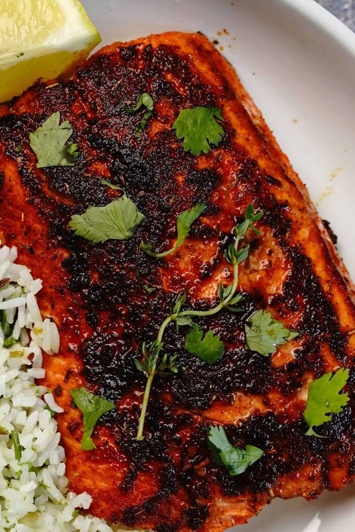 A piece of jerk salmon garnished with cilantro is served on a white plate next to herbed rice and a lemon wedge.
