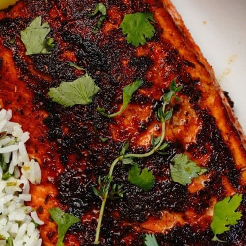 Jerk Salmon fillet with a dark seasoning crust, garnished with fresh herbs, served beside white rice and a lemon wedge on a white plate.