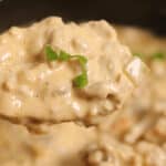 Close-up of a spoonful of creamy Philly Cheesesteak Dip, adorned with chopped green onions, poised over a larger pot filled with the same savory goodness.