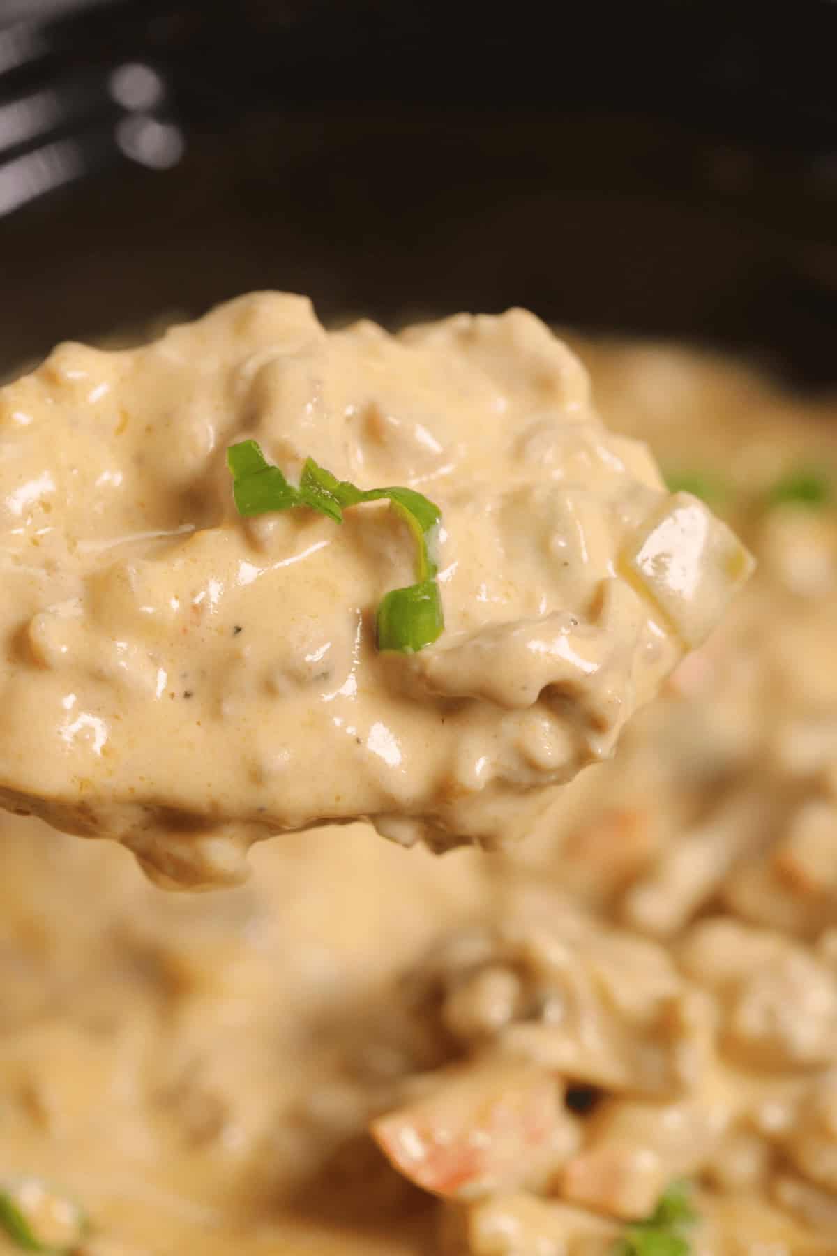 Close-up of a spoonful of creamy Philly Cheesesteak Dip, adorned with chopped green onions, poised over a larger pot filled with the same savory goodness.
