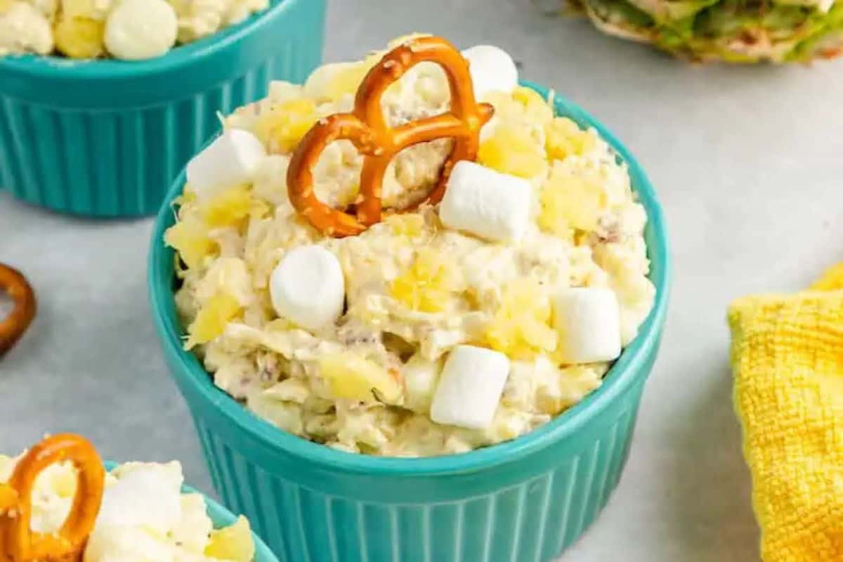 A teal bowl brimming with a creamy pineapple fluff, crowned with mini marshmallows and a pretzel, offers a delightful Easter dessert for kids. Nearby, another bowl and a yellow cloth peek into view, completing the festive scene.