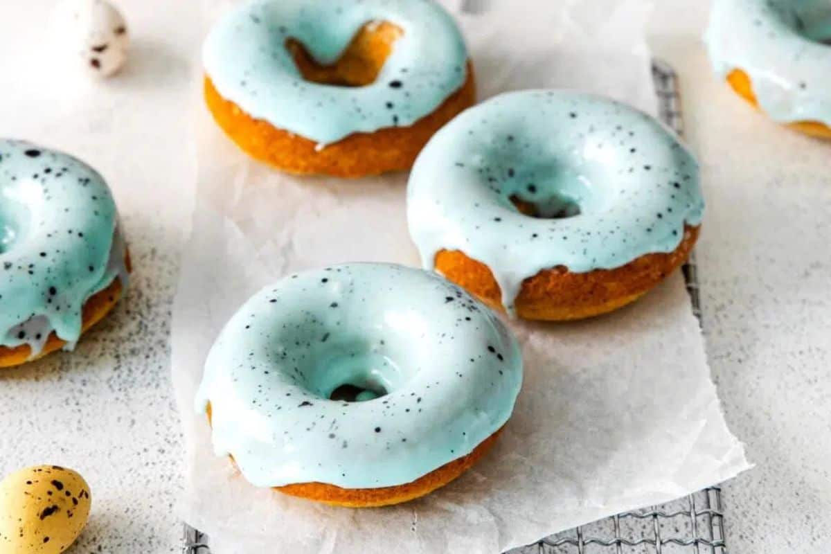 Three donuts with light blue icing and speckles sit on parchment paper, perfect Easter treats for kids, with a small egg-shaped item nearby.