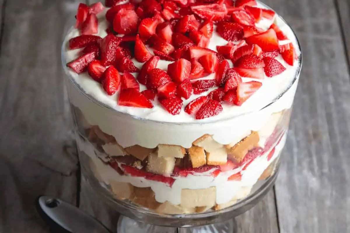 A delightful Easter dessert for kids, this layered strawberry trifle sits on a wooden surface, featuring sliced strawberries, cream, and sponge cake. A metal spoon lies invitingly beside the glass dish.