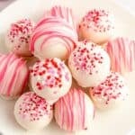 A plate of white chocolate-covered strawberry truffles decorated with pink and red sprinkles and a pink icing drizzle.