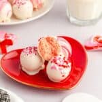 Three white chocolate-coated treats with pink and red sprinkles sit on a red heart-shaped plate, one cut in half to reveal the inside. A glass of milk and more cake balls are in the background.