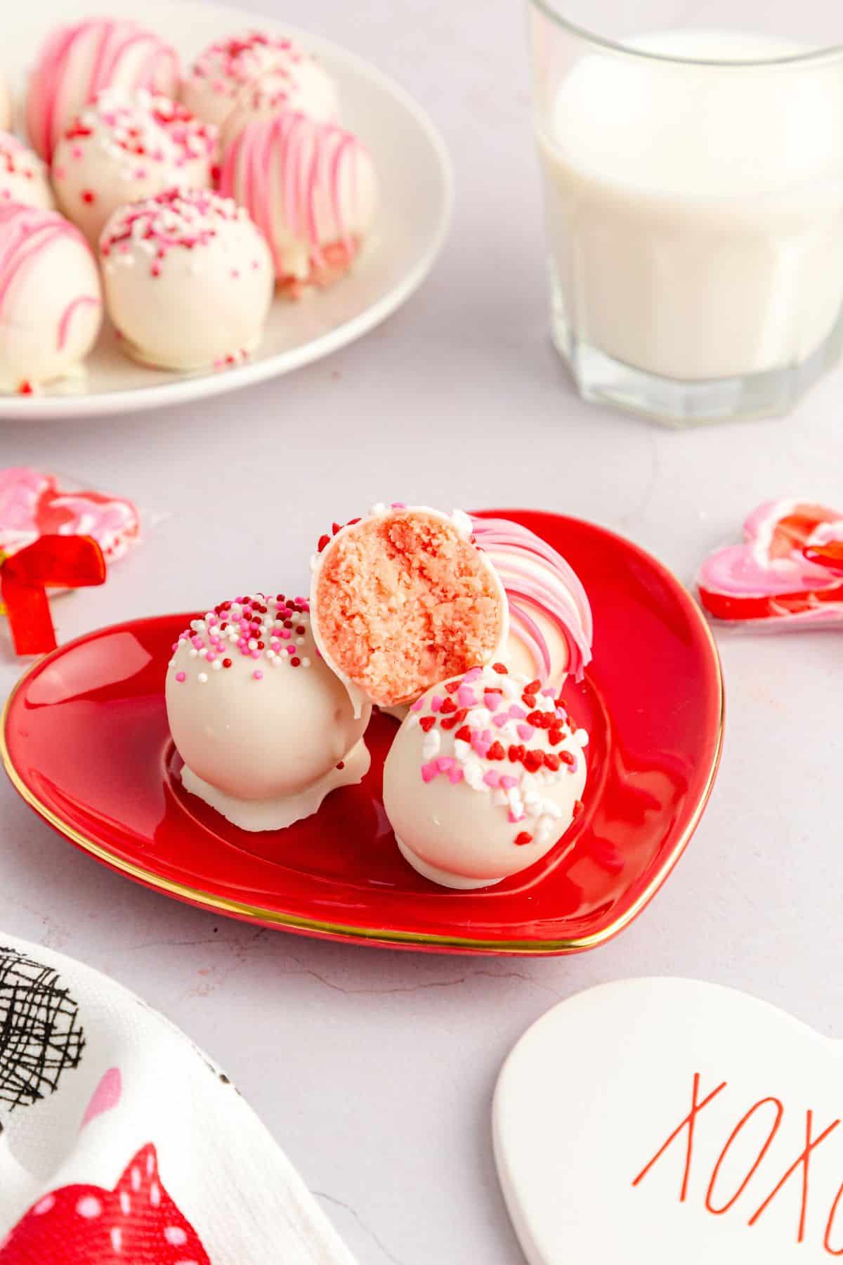 Three white chocolate-coated treats with pink and red sprinkles sit on a red heart-shaped plate, one cut in half to reveal the inside. A glass of milk and more cake balls are in the background.