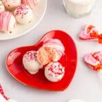 A red heart-shaped plate holds white chocolate cake balls with pink sprinkles, resembling strawberry truffles, one cut in half, next to a glass of milk and heart-shaped candies.