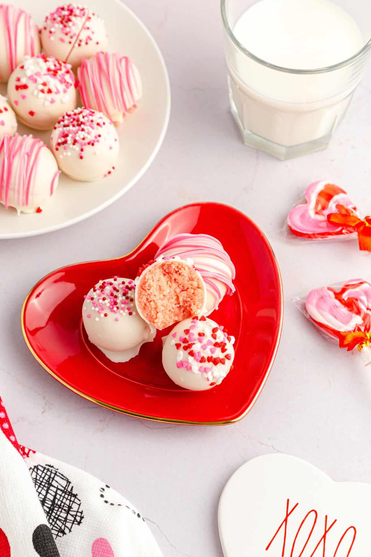 A red heart-shaped plate holds white chocolate cake balls with pink sprinkles, one cut in half, next to a glass of milk and heart-shaped candies.