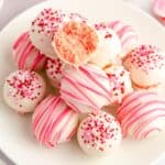 A plate of white chocolate-coated strawberry truffles decorated with pink and red sprinkles and stripes, with one truffle cut open to reveal a pink interior.