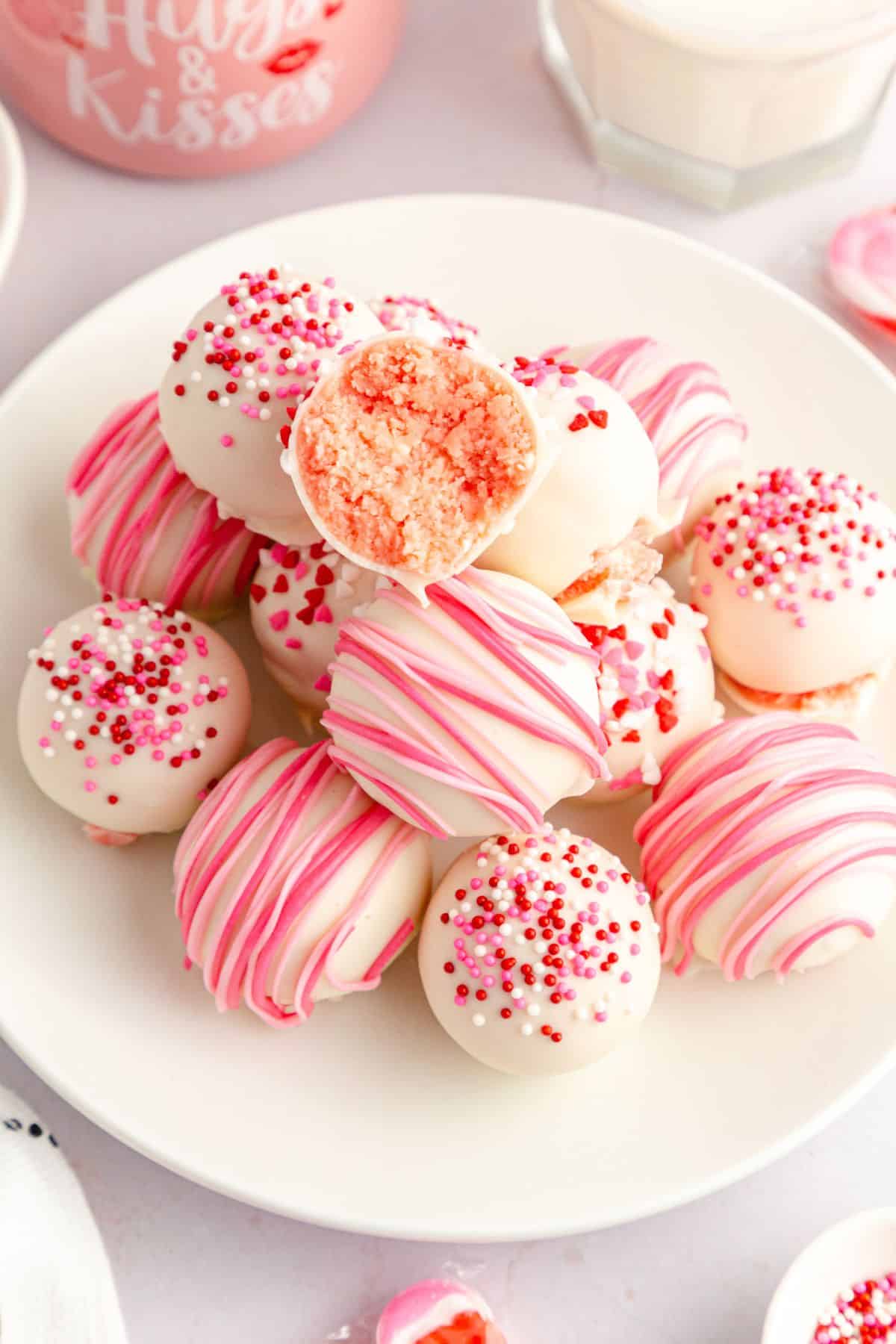 A plate of white chocolate-coated strawberry truffles decorated with pink and red sprinkles and stripes, with one truffle cut open to reveal a pink interior.