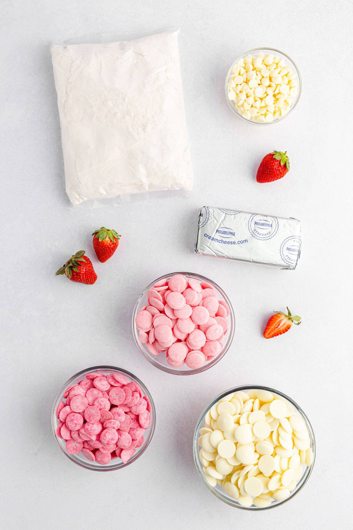 Bowls of pink and white candy melts, white chocolate chips, cream cheese, powdered sugar, and fresh strawberries are arranged on a white surface.