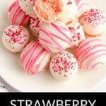 A plate of Strawberry Truffles coated in white chocolate, some adorned with pink stripes and sprinkles, with one truffle cut open to reveal a luscious pink center. Text reads "Strawberry Truffles.