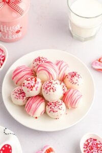 A plate of strawberry truffles coated in white chocolate, decorated with pink drizzle and red, pink, and white sprinkles, sits next to a glass of milk and Valentine's-themed items.