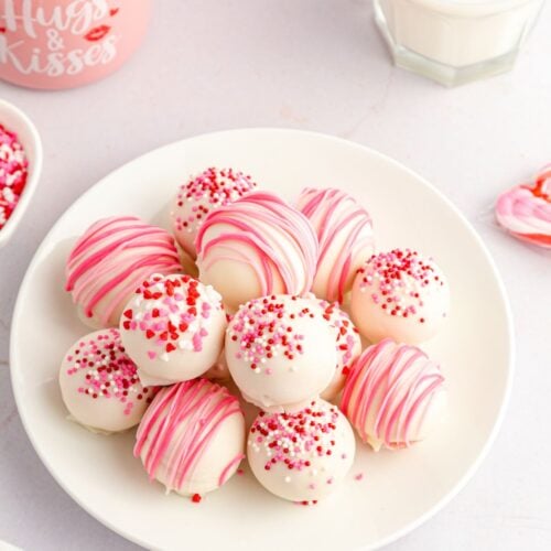 A plate of strawberry truffles coated in white chocolate, decorated with pink drizzle and red, pink, and white sprinkles, sits next to a glass of milk and Valentine's-themed items.