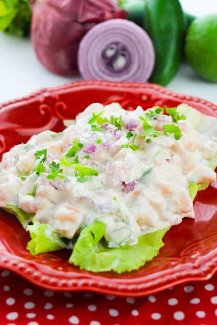 A creamy chicken salad with chopped vegetables is served on lettuce leaves and garnished with fresh herbs, presented on a red plate. Reminiscent of a classic Shrimp Salad Sandwich, sliced onion, jalapeño, and lime are arranged in the background for a burst of color and flavor.
