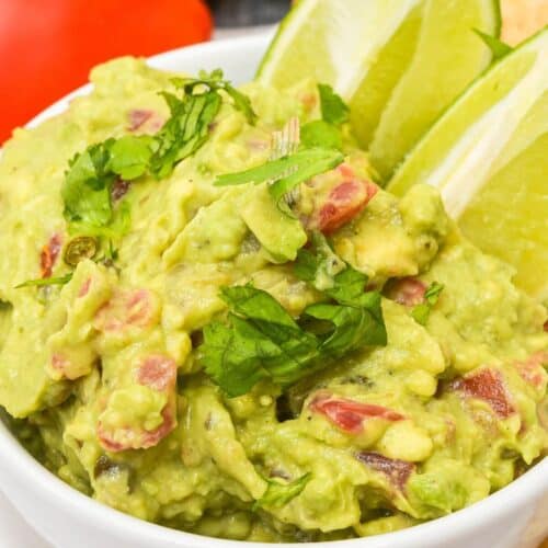 A bowl of 4-ingredient guacamole recipe is garnished with cilantro and served with lime wedges, with tomatoes visible in the background.