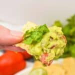 A hand holds a tortilla chip topped ingredient recipe and fresh cilantro, with tomatoes, lime wedges, and more chips in the background.
