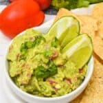 A bowl of 4-ingredient guacamole recipe topped with chopped cilantro and lime wedges, surrounded by tortilla chips, tomatoes, and fresh cilantro.