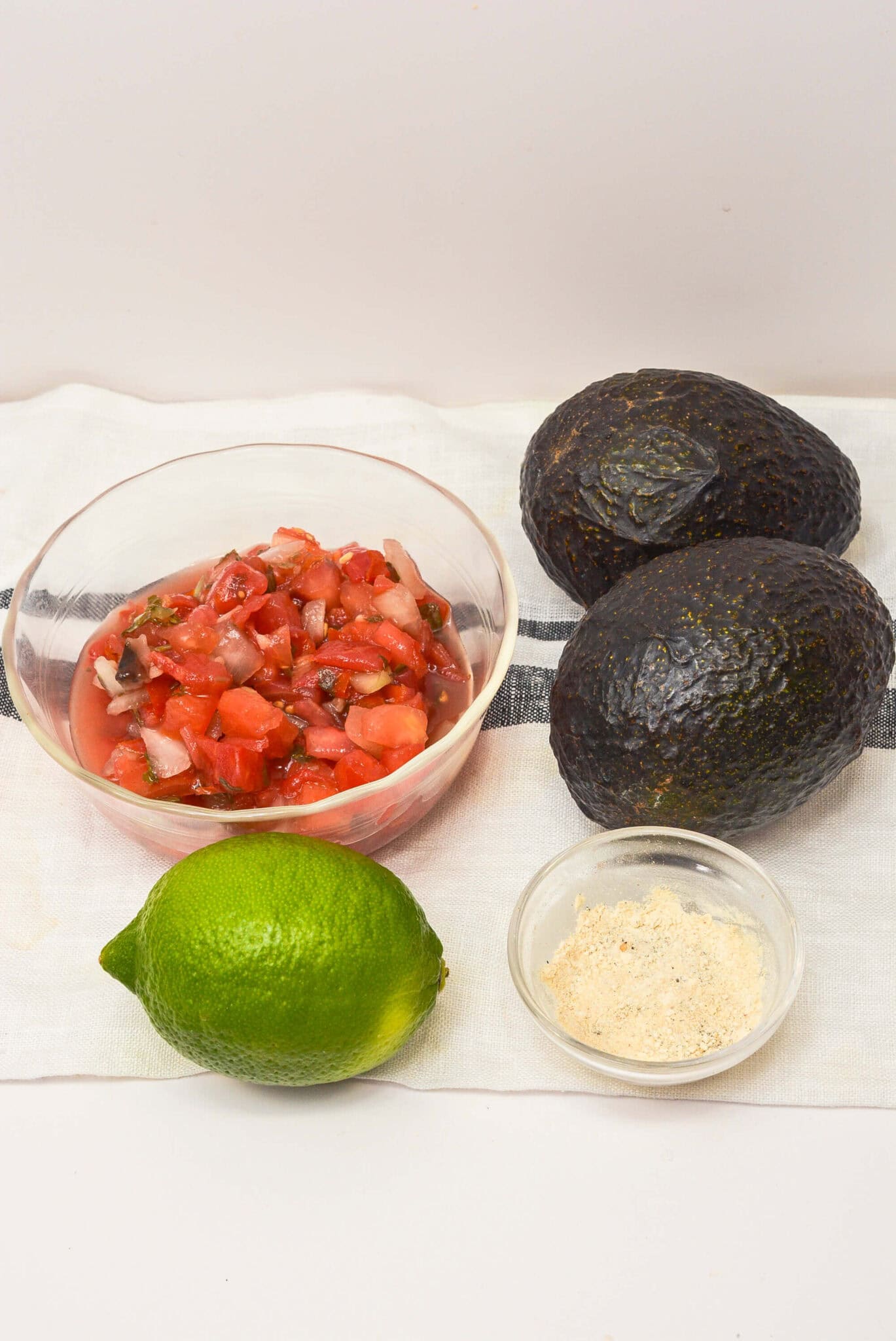 A bowl of salsa, two whole avocados, a lime, and a small dish of seasoning are arranged on a white cloth—perfect ingredients.