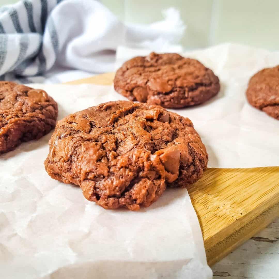 Four Brownie Cake Cookies from mix, with visible chocolate chunks, are placed on parchment paper atop a wooden board, with a striped cloth in the background.