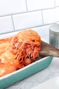 A wooden spatula lifts a serving of cheesy spaghetti with melted cheese from a turquoise casserole dish on a kitchen counter.